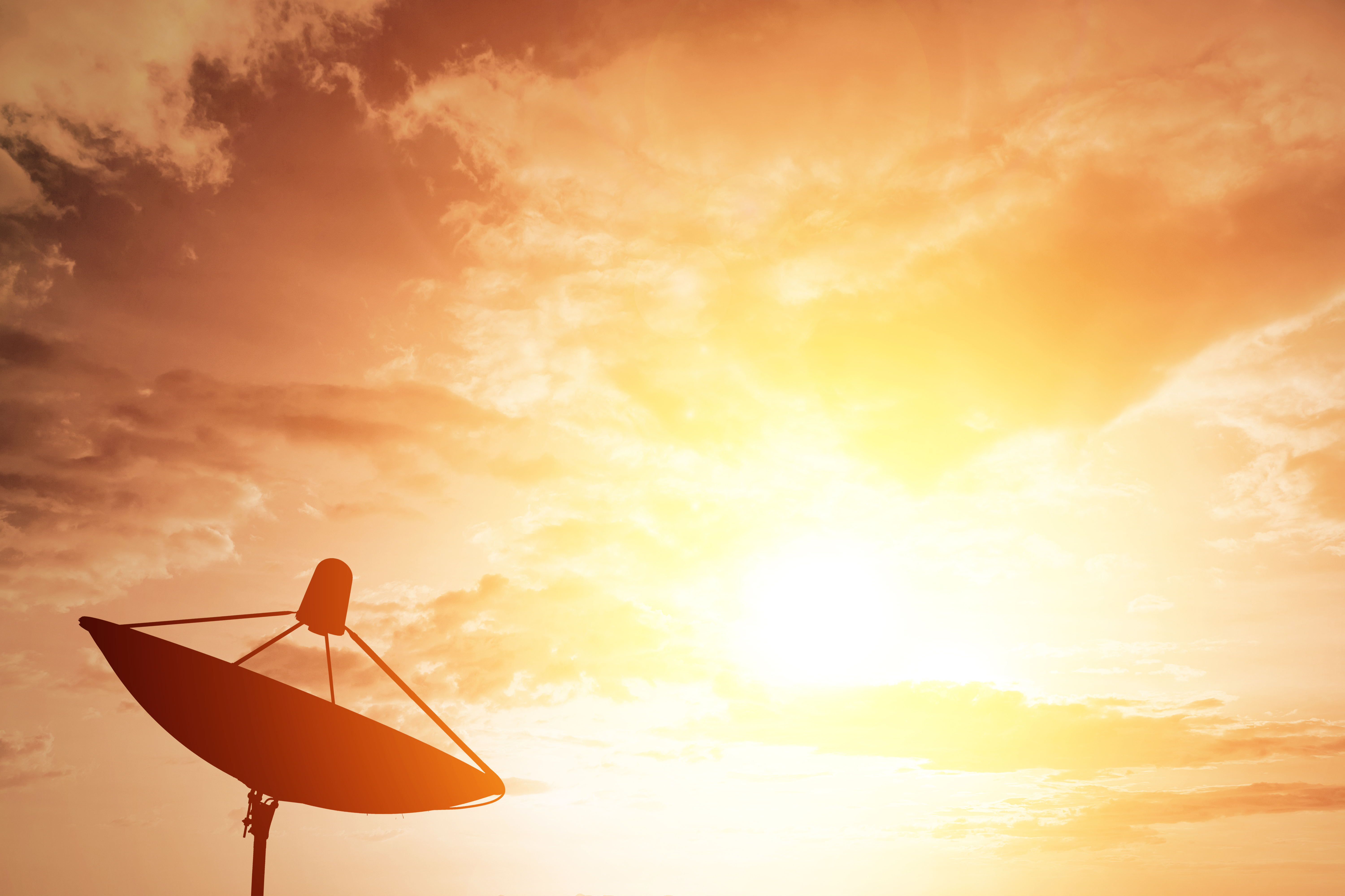 Large satellite dish silhouetted against a bright sunburst sky during sunset, symbolizing satellite signal interference from sunspot activity.
